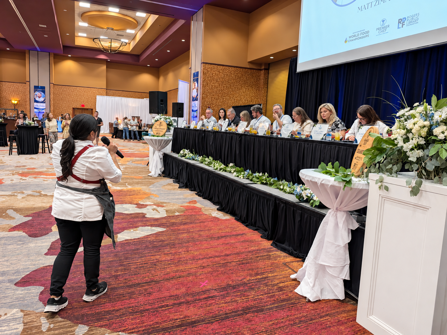 A chef speaks into a microphone in front of the Premier Chef Northwest Arkansas judges